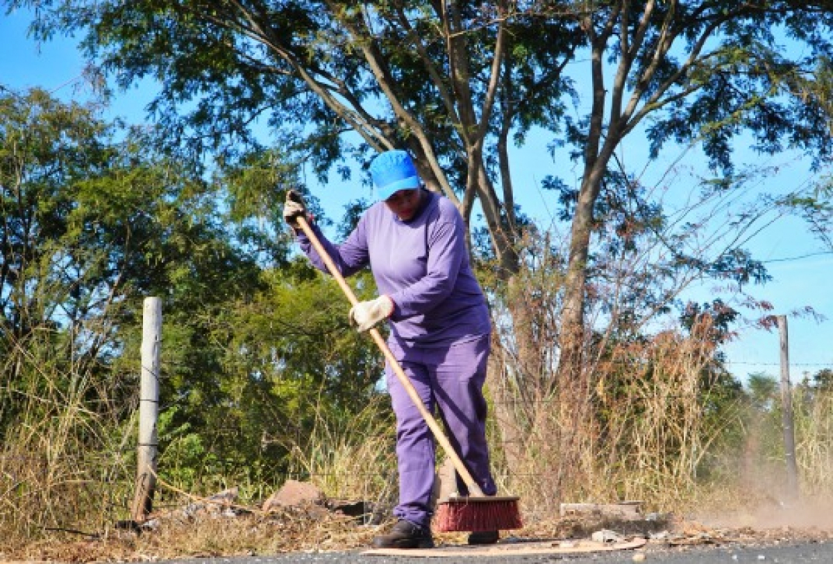 Servidora realiza varrição e limpeza às margens de uma via pública em área rural.