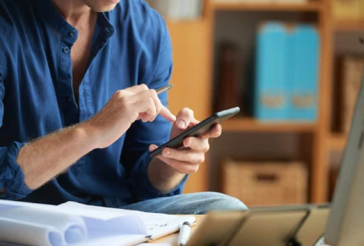 Pessoa utilizando um celular enquanto trabalha em uma mesa com papéis e computador.