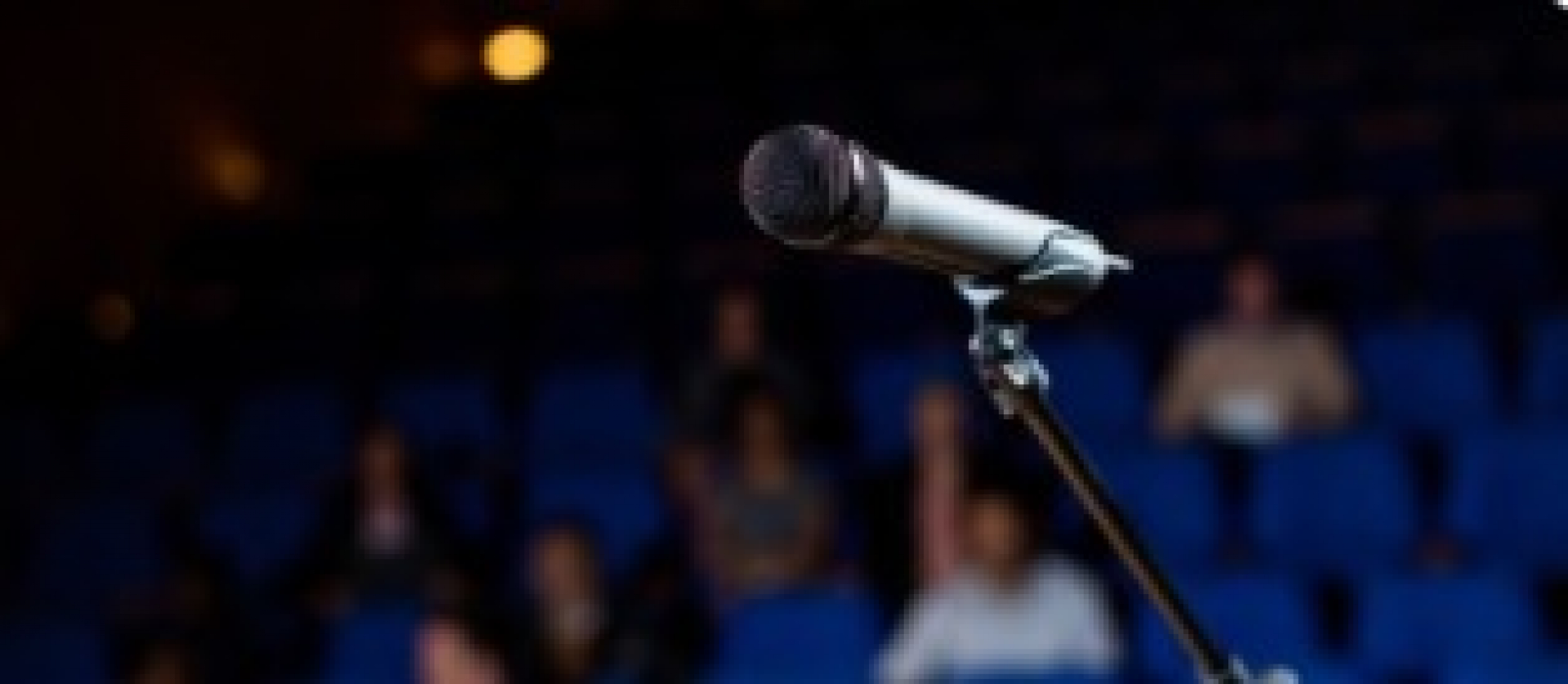 Microfone em destaque em um auditório com plateia ao fundo, pronto para uma apresentação.