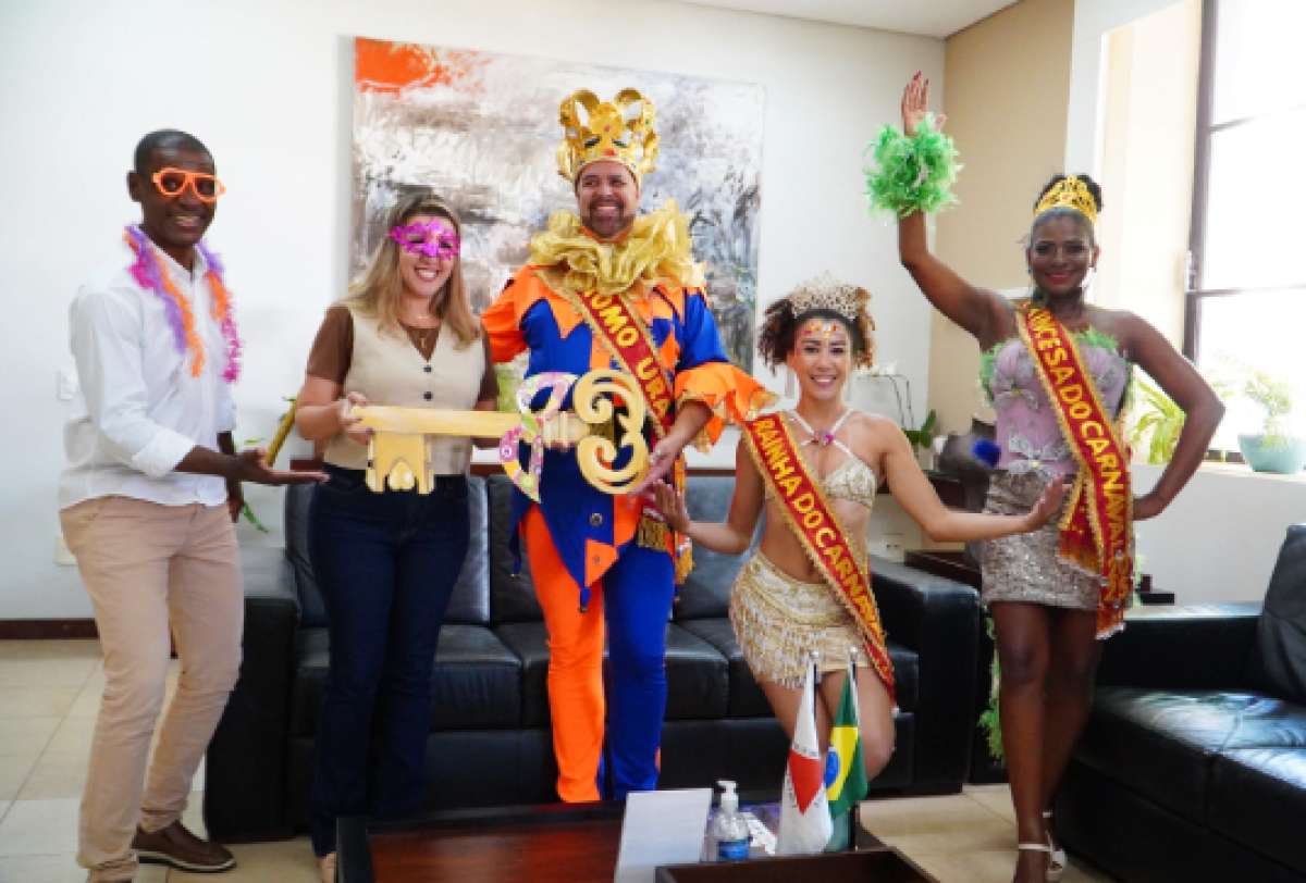 Uma vibrante celebração de carnaval com indivíduos em trajes coloridos, incluindo uma Embaixadora e uma Rainha do Carnaval.