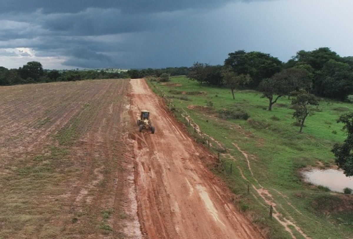 A imagem mostra uma máquina niveladora trabalhando na abertura ou manutenção de uma estrada de terra em área rural.