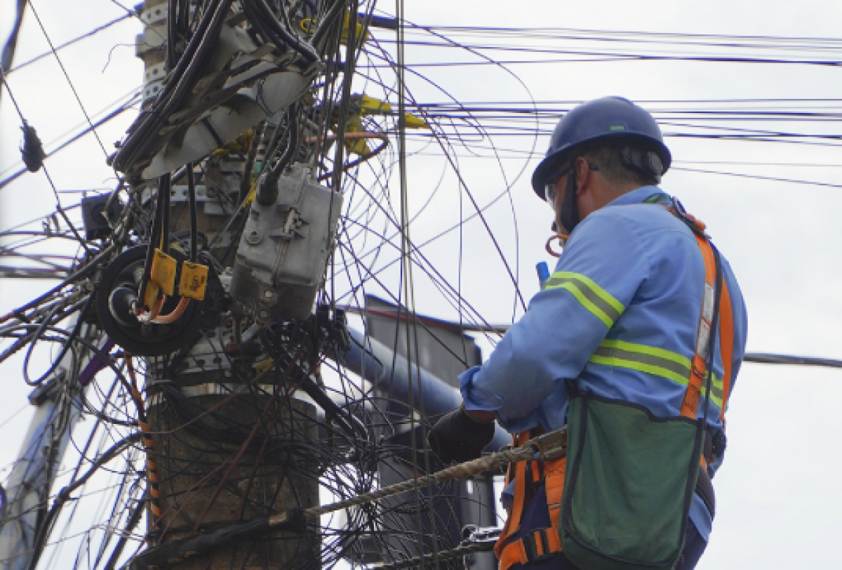Trabalhador realiza manutenção em um poste repleto de fios emaranhados, utilizando equipamentos de segurança.