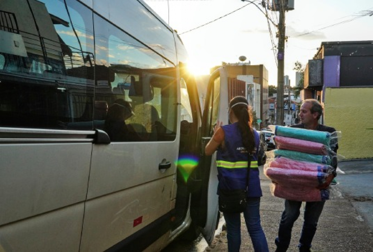 Profissionais realizam entrega de cobertores ao lado de uma van, em ação social durante o entardecer em área urbana.