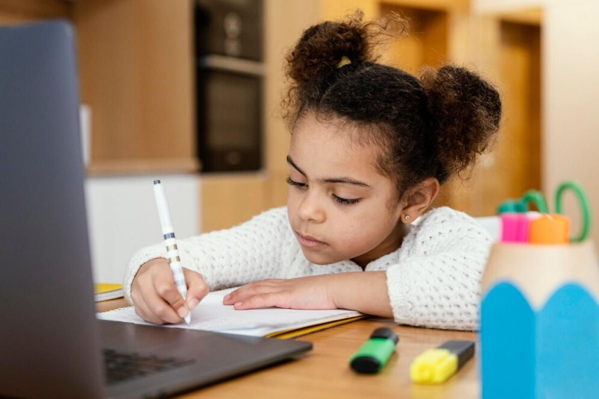 Menina concentrada escreve em caderno enquanto acompanha algo no notebook, em ambiente doméstico.