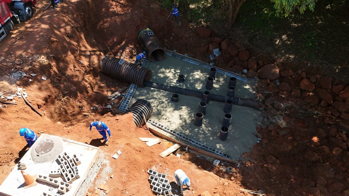 Obra de infraestrutura em andamento, com trabalhadores instalando tubulações e estruturas de concreto em uma área escavada