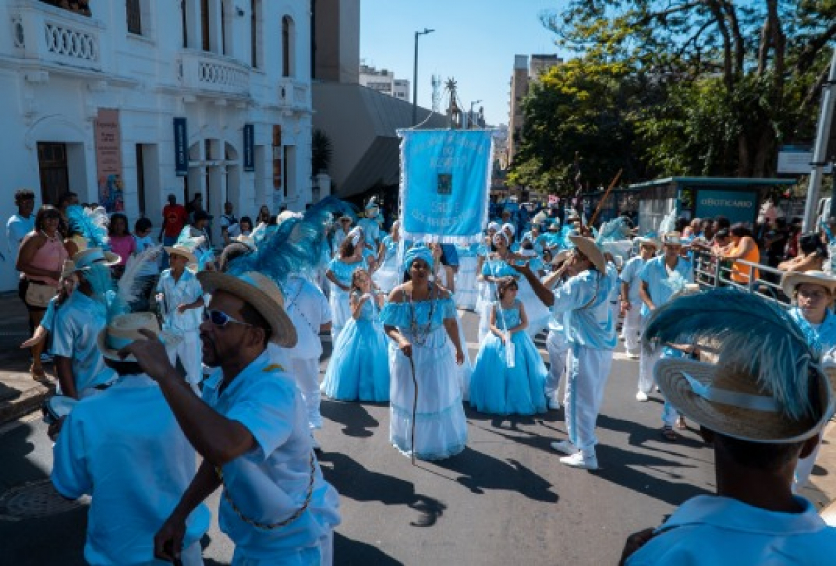 A imagem mostra um grupo folclórico participando de um desfile ou procissão cultural. 