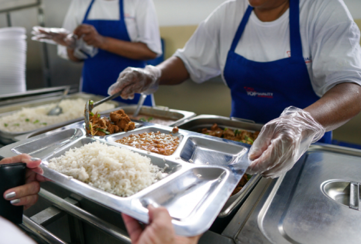 Distribuição de refeições em um restaurante popular, com atendentes servindo comida em bandejas de aço inox.