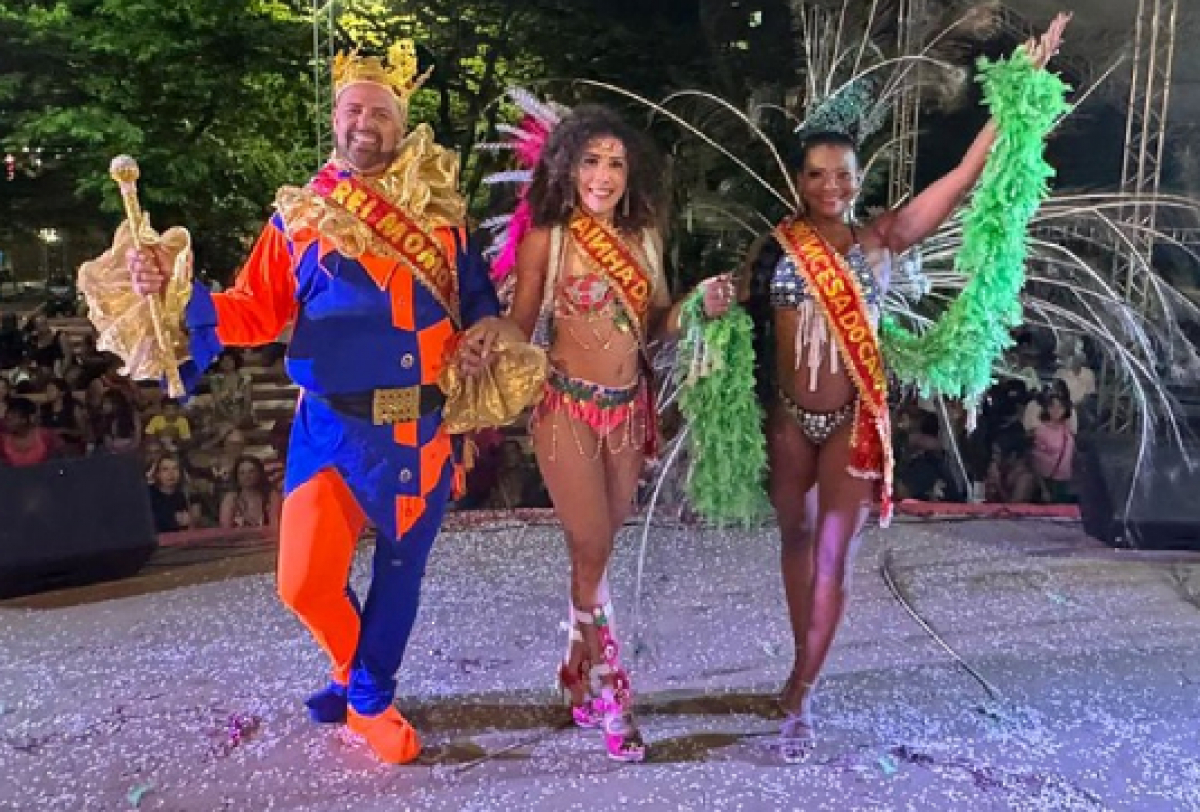 Trio festivo em um evento de carnaval, com trajes coloridos e faixas de destaque.