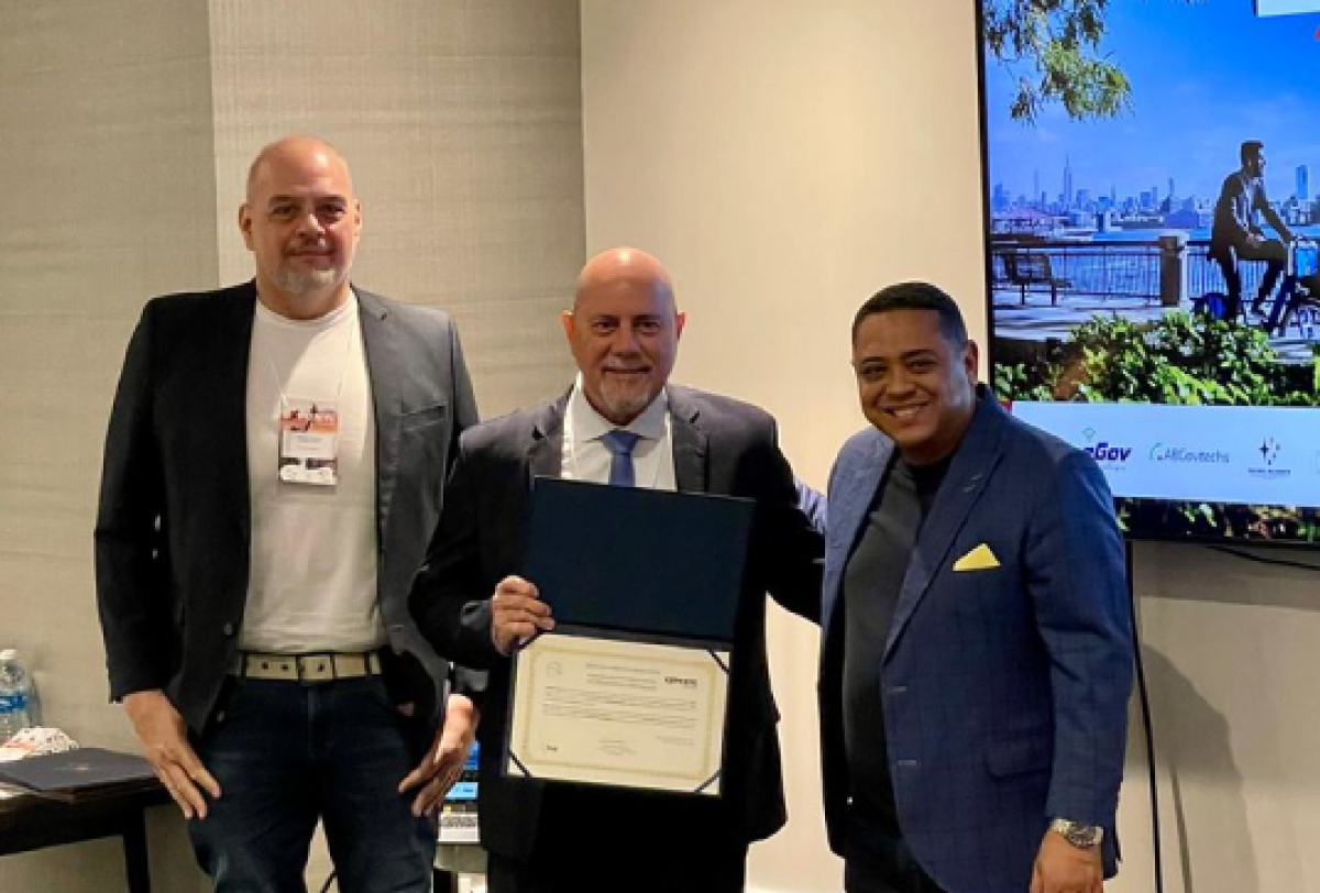 Três homens posam para uma foto, com um deles segurando um certificado, em um ambiente corporativo.