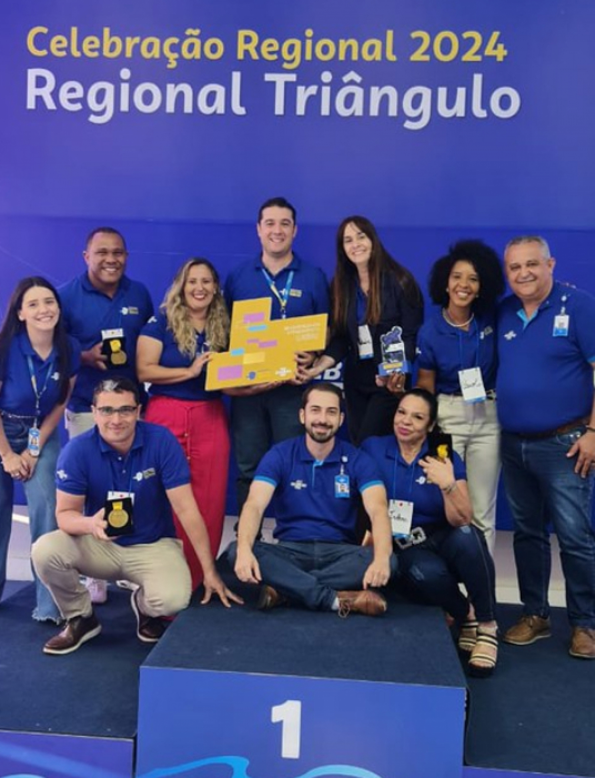 A imagem mostra um grupo de pessoas posando juntas em um pódio com um painel de fundo que lê Celebração Regional 2024 Regional Triângulo. 