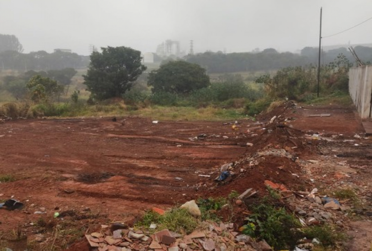 Terreno com entulho e resíduos em área aberta, cercado por vegetação e com clima nublado.