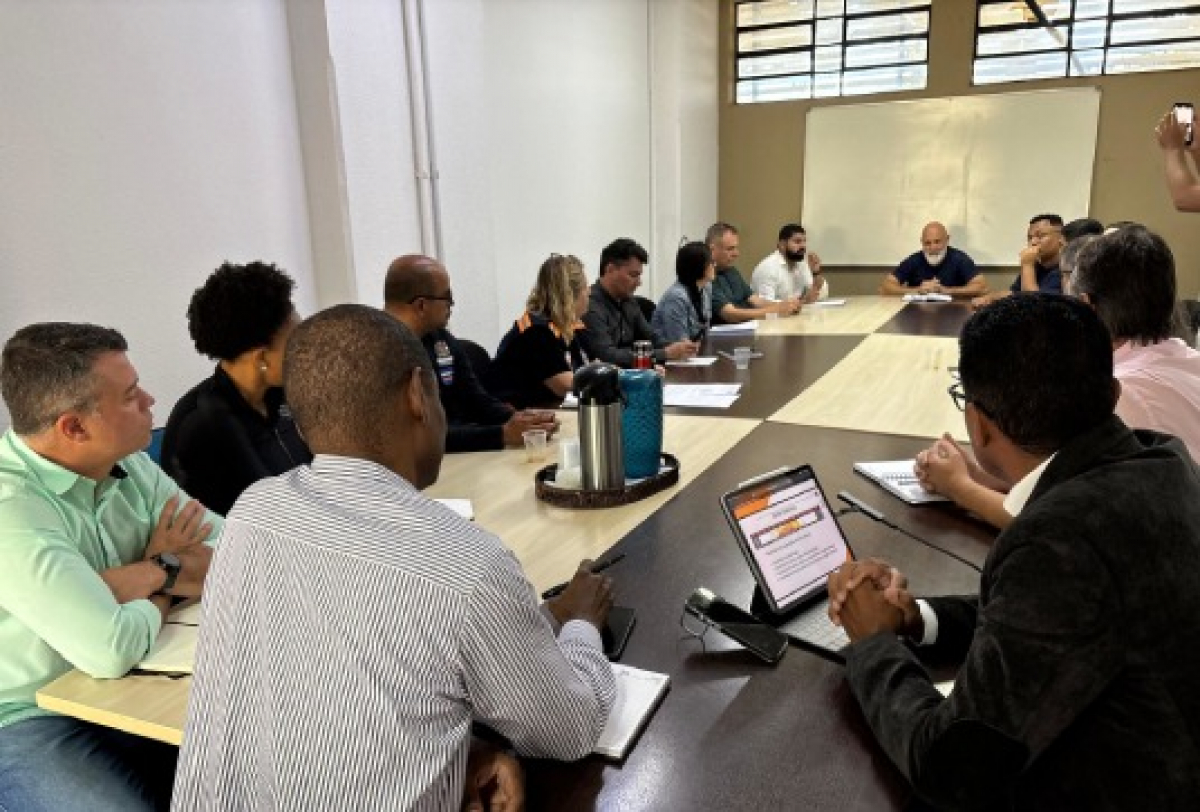 A imagem mostra uma reunião de trabalho com várias pessoas sentadas ao redor de uma mesa discutindo assuntos, com notebooks.