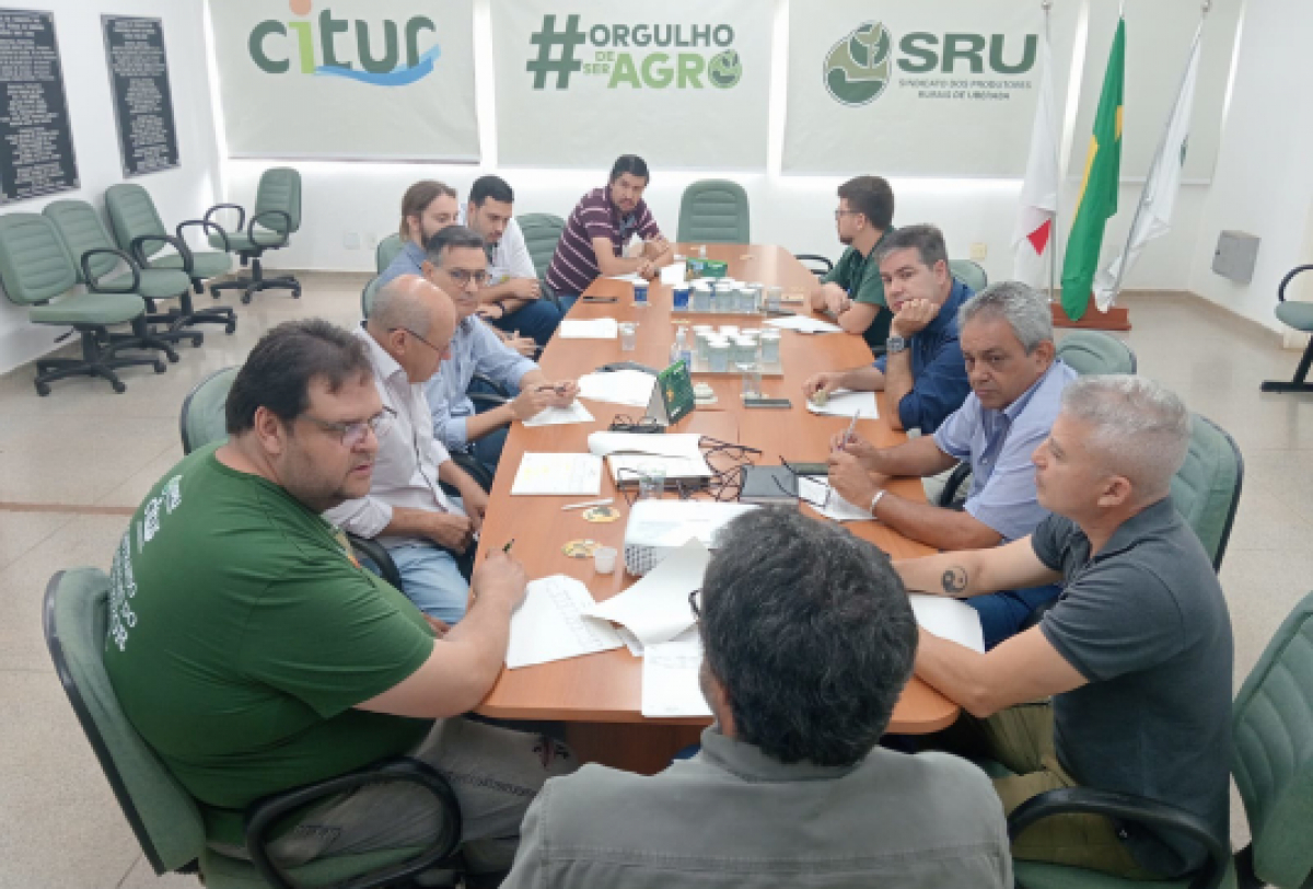 A imagem mostra um grupo de pessoas sentadas em torno de uma grande mesa de conferência em uma sala de reuniões, engajadas em uma discussão. 