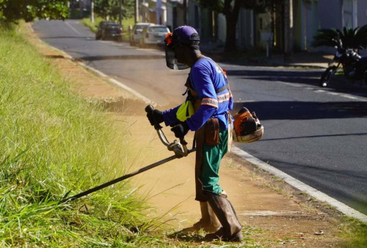 Trabalhador aparando grama com segurança ao longo da calçada.