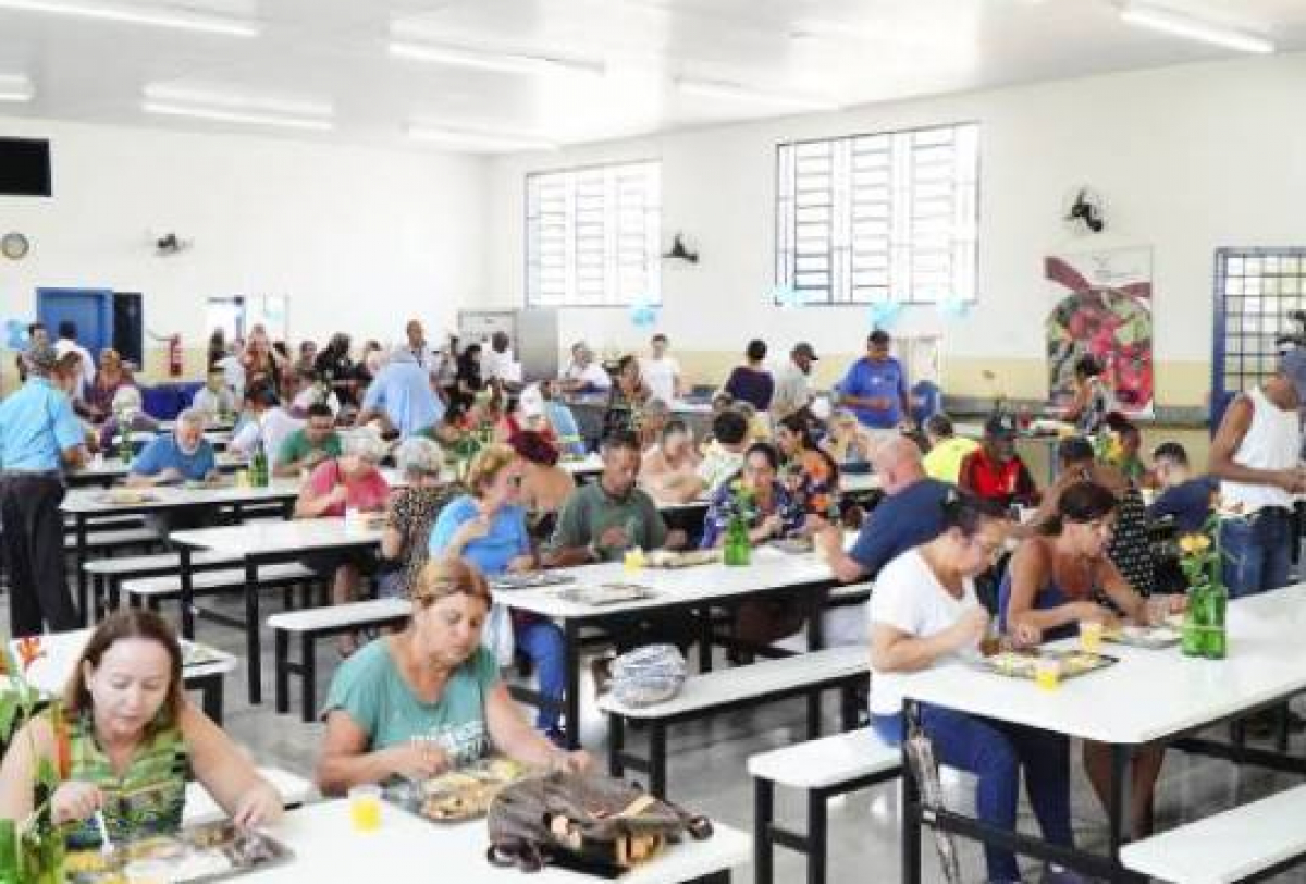 Pessoas almoçam em um restaurante popular movimentado, com ambiente iluminado e organizado.       Você disse: