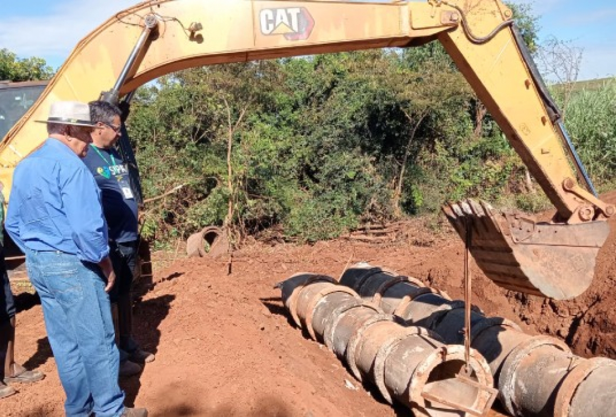 Supervisores acompanham a instalação de tubos de concreto com auxílio de uma escavadeira em obra de drenagem.
