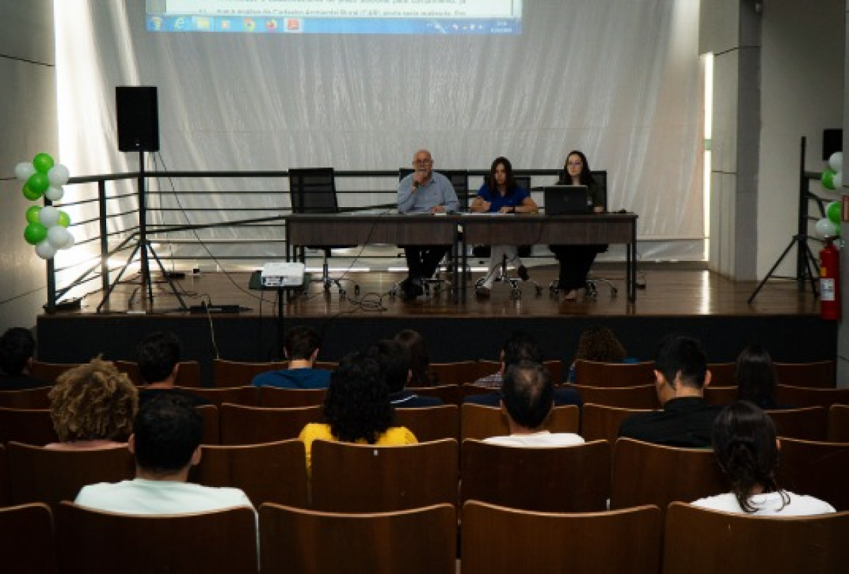 A imagem mostra um evento ou palestra com um painel de três pessoas à frente, enquanto o público, em cadeiras, assiste atentamente.