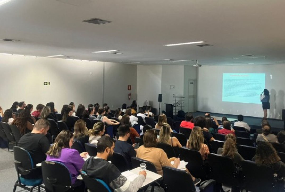 Palestra em auditório com grande público atento à apresentação de uma mulher, que utiliza slides projetados na tela.