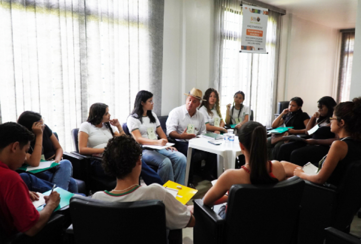 A imagem mostra um grupo de pessoas sentadas em um círculo em uma sala, participando de uma discussão ou reunião. 