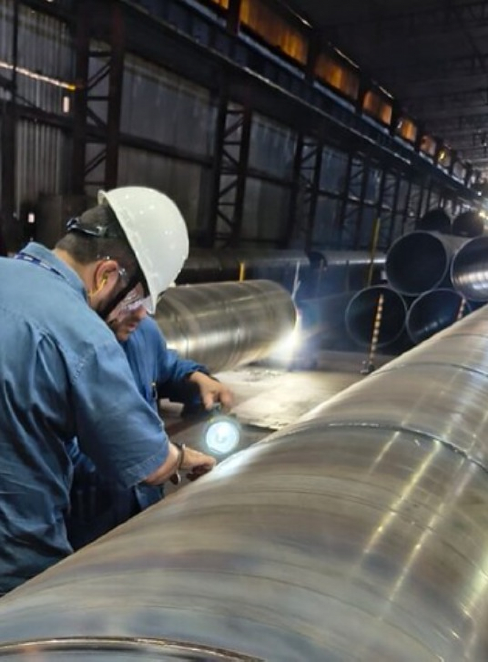Funcionários com capacetes e equipamentos de segurança inspecionam grandes tubos metálicos em uma indústria.