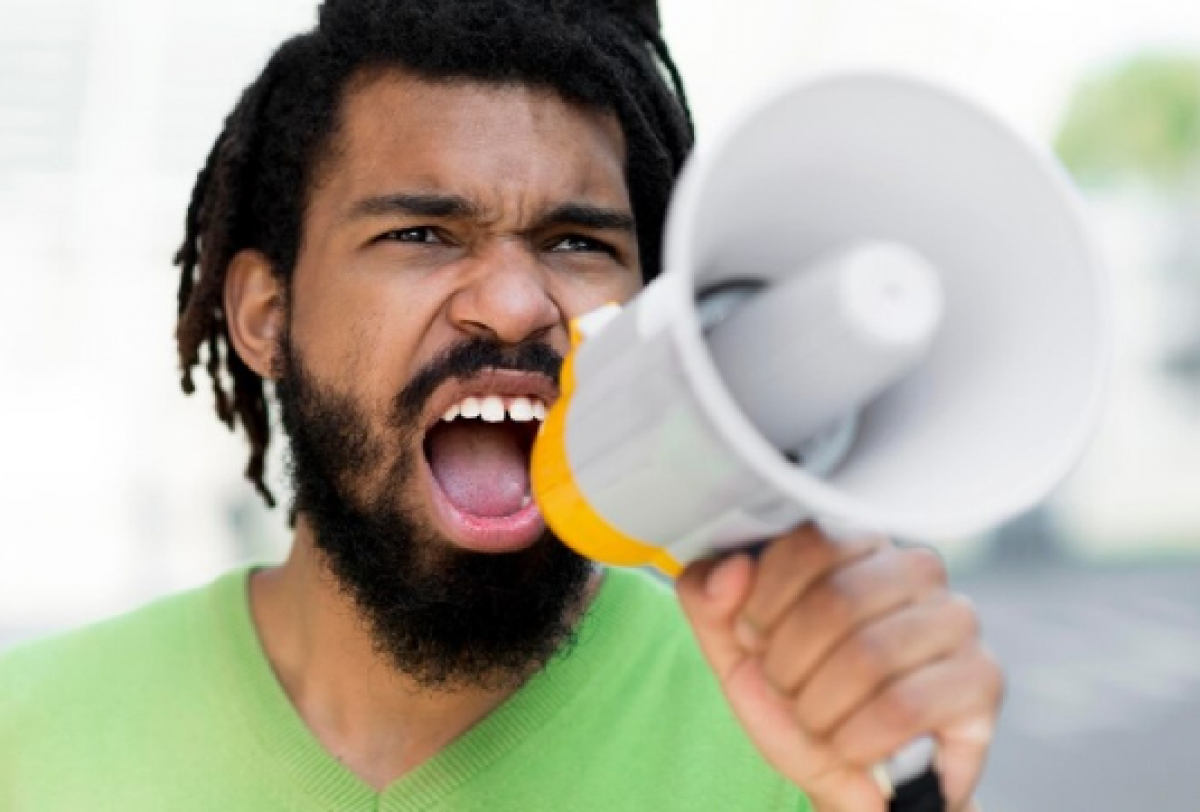 A imagem mostra uma pessoa segurando um megafone em um ambiente ao ar livre, possivelmente fazendo um anúncio ou discurso. 