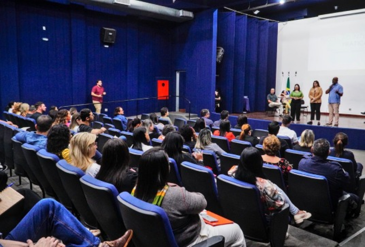 Imagem mostra um grupo de pessoas participando do que parece ser uma apresentação formal ou seminário em um auditório.