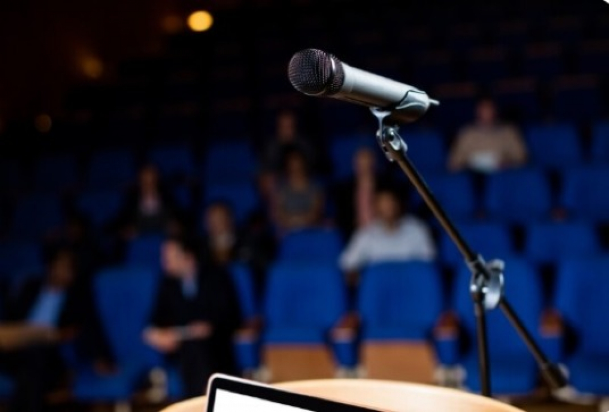 Microfone em destaque em um auditório com plateia ao fundo, pronto para uma apresentação.