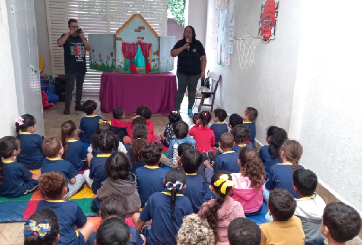 Apresentadores realizam uma contação de histórias com teatro de fantoches para um grupo de crianças em uma escola.
