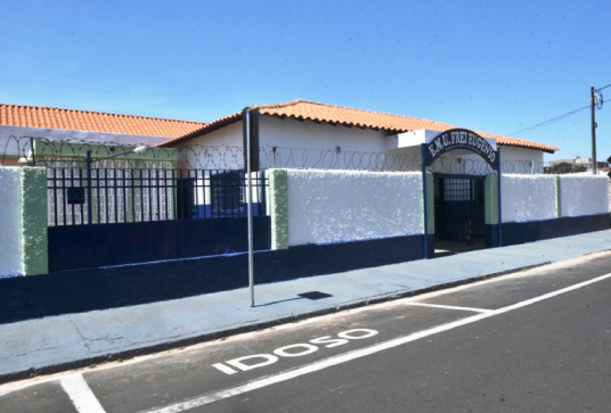 Fachada da Escola Municipal Frei Eugênio, com muros brancos e azuis, portões metálicos e vaga de estacionamento reservada para idosos.