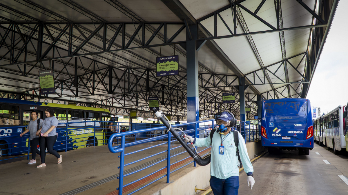 A imagem mostra uma estação de transporte público, com estrutura metálica coberta e diversas plataformas sinalizadas. Um trabalhador com equipamento de proteção individual (EPI), incluindo máscara, luvas e óculos de segurança, está realizando a desinfecção do local utilizando um pulverizador. No plano de fundo, há ônibus azuis estacionados, com detalhes como logotipos e números de identificação visíveis. Duas pessoas estão caminhando pela área da plataforma, que possui corrimões azuis e acessibilidade.