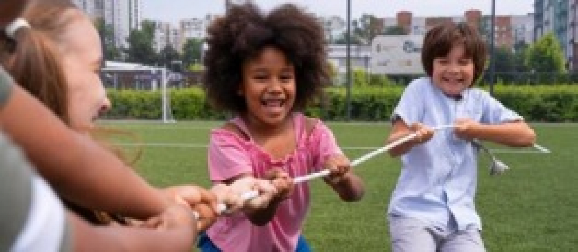 A imagem mostra um grupo de crianças jogando cabo de guerra em um campo de grama, com um fundo de campo esportivo.