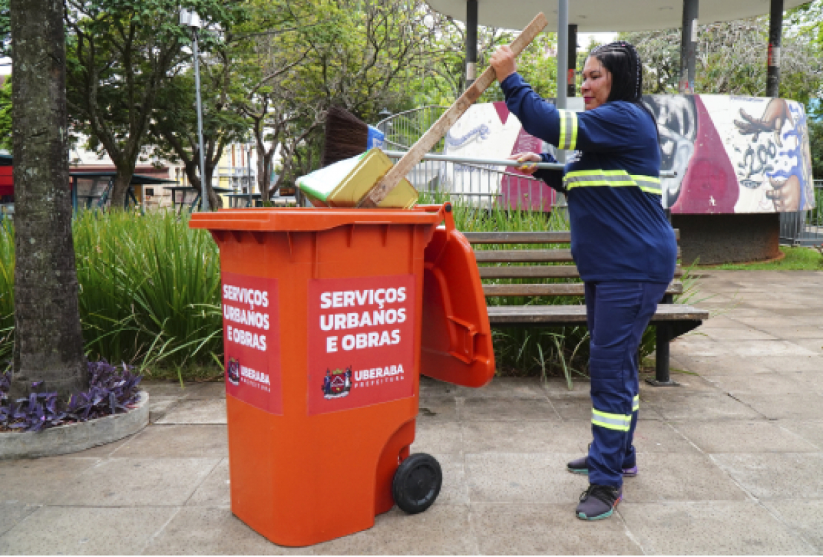 A imagem mostra uma pessoa em uniforme azul descartando lixo em uma lixeira laranja, realizando serviços de limpeza pública em um parque.