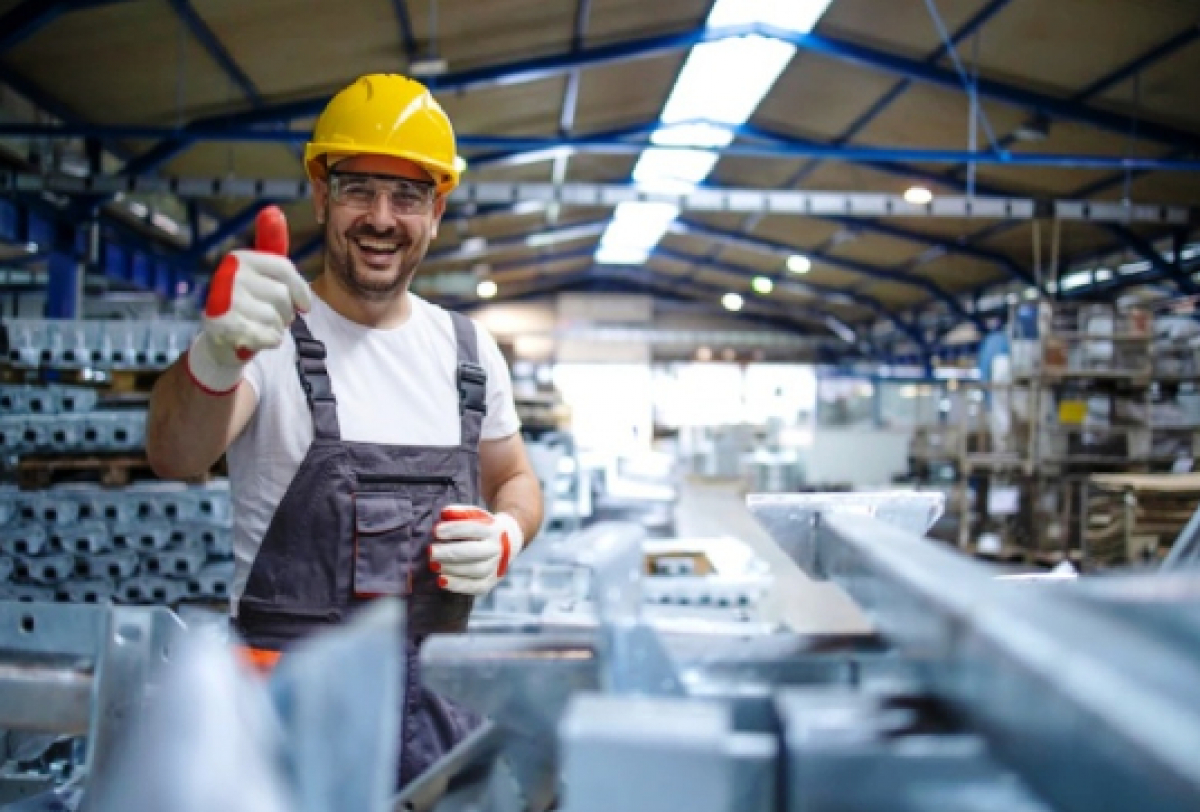 Trabalhador da construção civil, usando capacete e equipamentos de proteção, faz sinal de positivo dentro de uma fábrica.