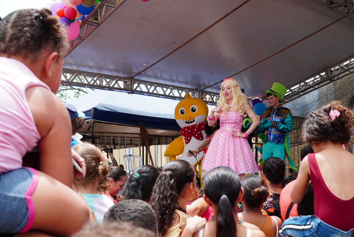 Crianças assistem animadas a um espetáculo infantil com personagens fantasiados em um palco decorado com balões coloridos.