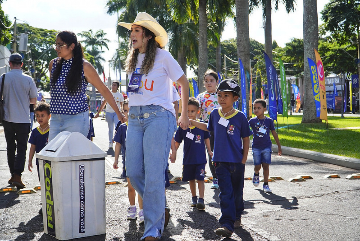 Mulher de chapéu conduz crianças uniformizadas por uma via arborizada durante uma atividade educativa.