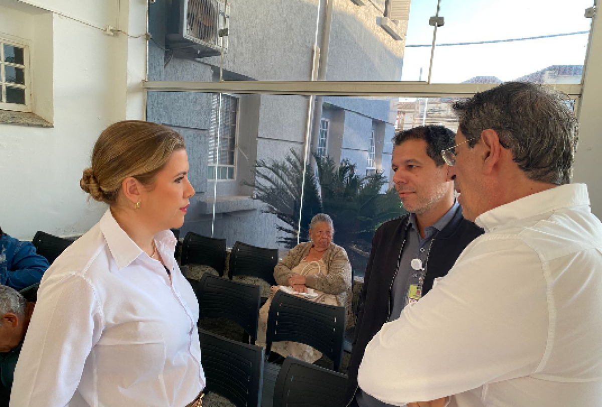 Autoridades conversam durante visita a unidade de saúde com presença de pacientes ao fundo. 