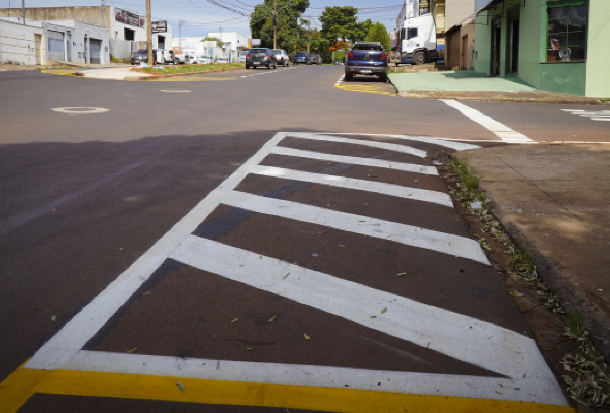 A imagem mostra uma rua com sinalização de faixas brancas diagonais entre linhas amarelas, indicando uma zona de proibição de estacionamento ou parada. 