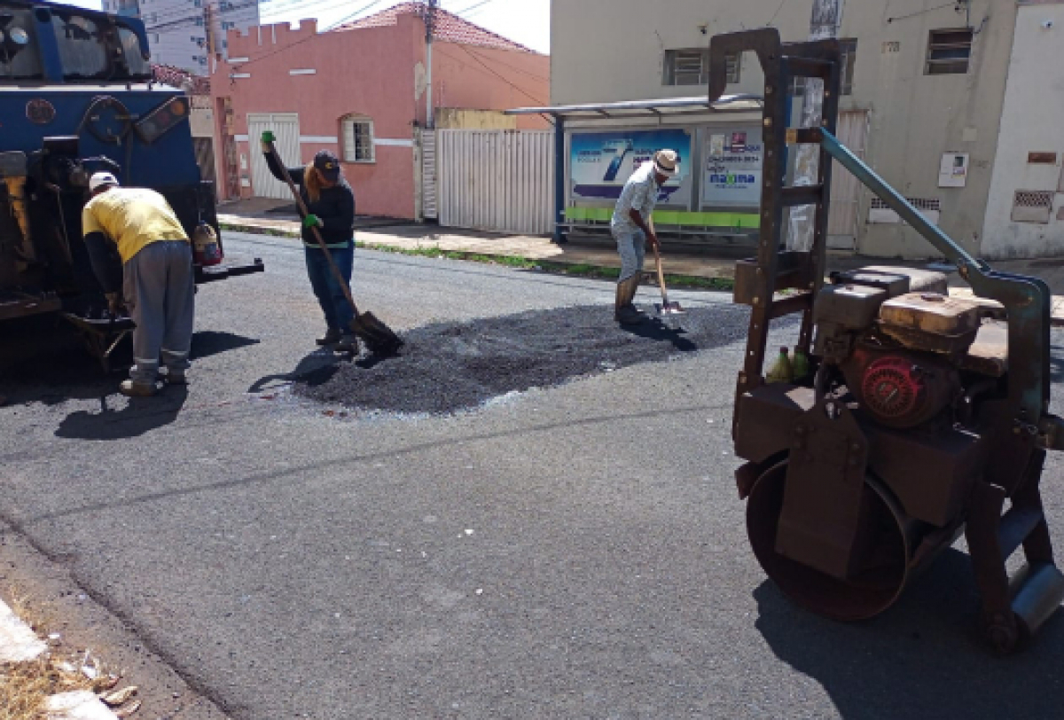 Equipe realiza reparo asfáltico com nivelamento manual e compactação com rolo mecânico.