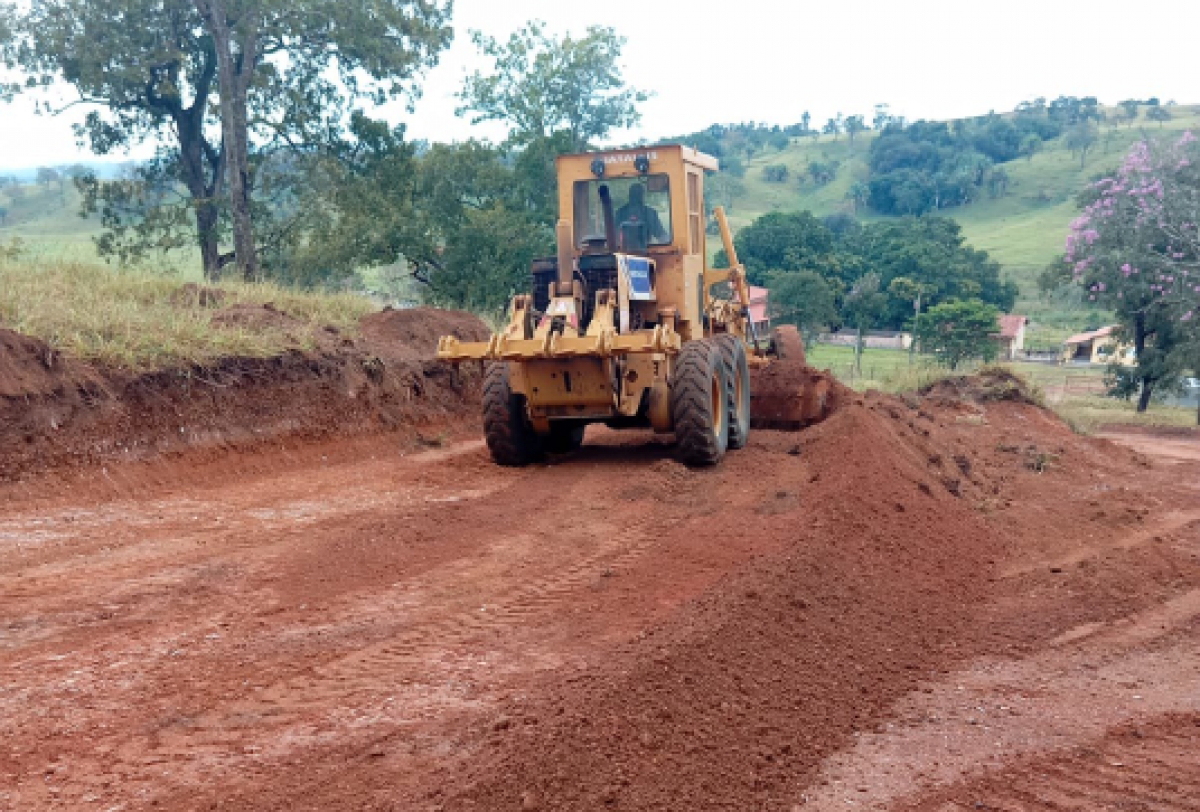 Uma máquina motoniveladora realiza a terraplanagem de uma estrada de terra em área rural.