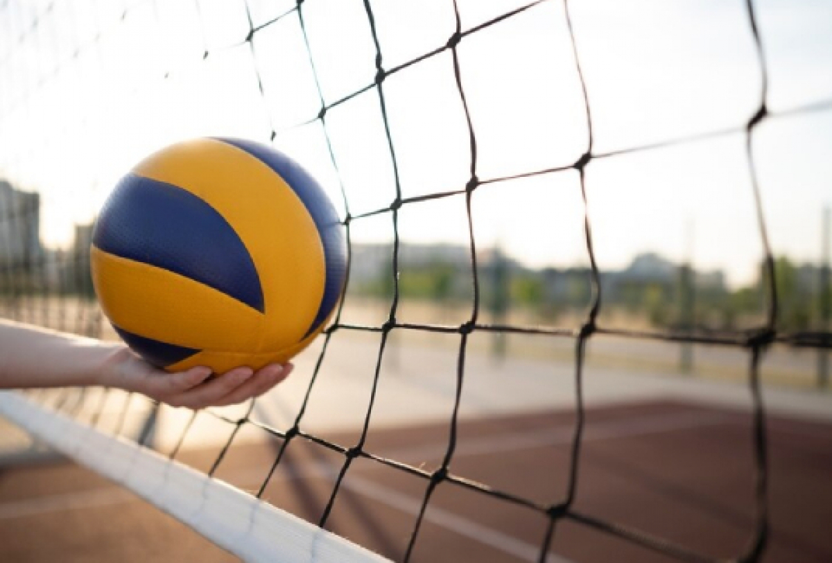 Quadra, uma mão segura uma bola de vôlei e a frente uma rede.