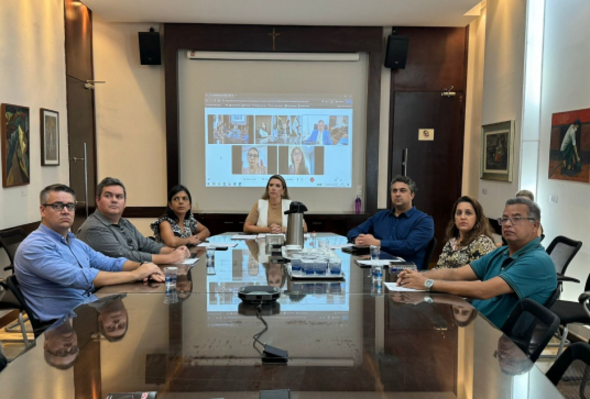 A imagem mostra um grupo de pessoas sentadas ao redor de uma grande mesa de conferência em uma sala de reuniões, com uma tela grande exibindo uma videoconferência ao fundo.