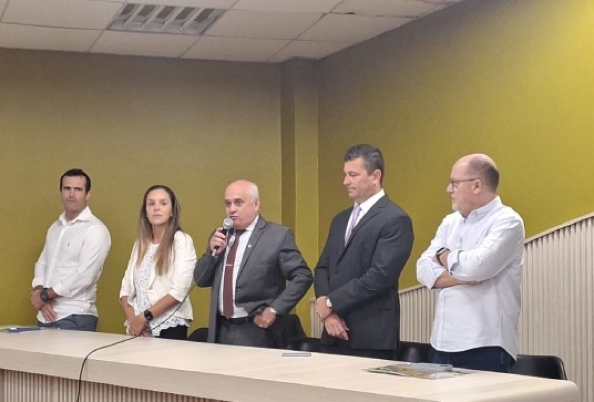 Cinco pessoas participam de um evento formal, com uma delas discursando ao microfone em uma sala iluminada.