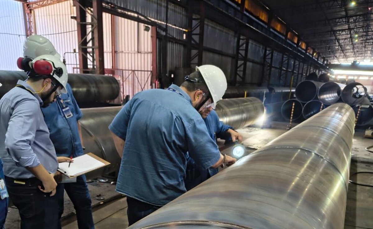 Funcionários com capacetes e equipamentos de segurança inspecionam grandes tubos metálicos em uma indústria.