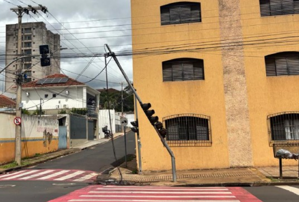 Semáforo inclinado em cruzamento urbano, com fiação exposta, indicando possível dano ou risco na via.