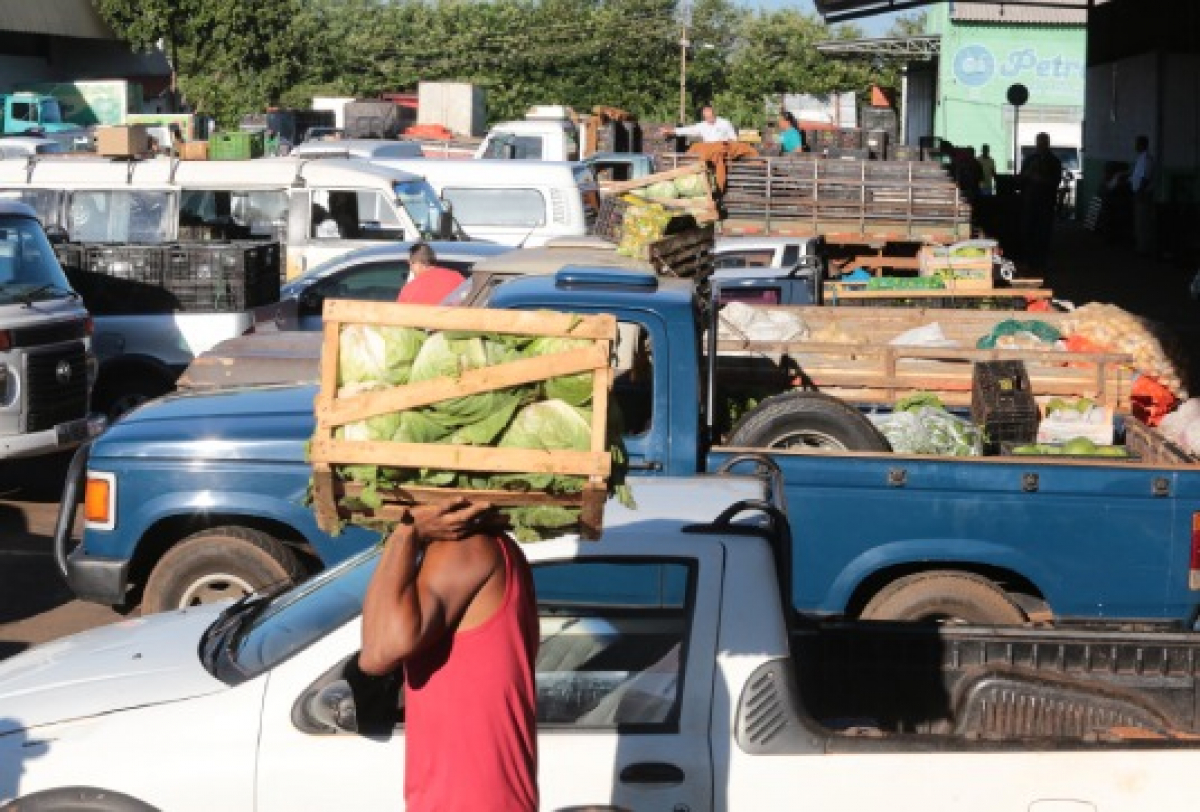 Centro de abastecimento com caminhonetes carregadas de hortaliças e trabalhador carregando caixa de verduras.