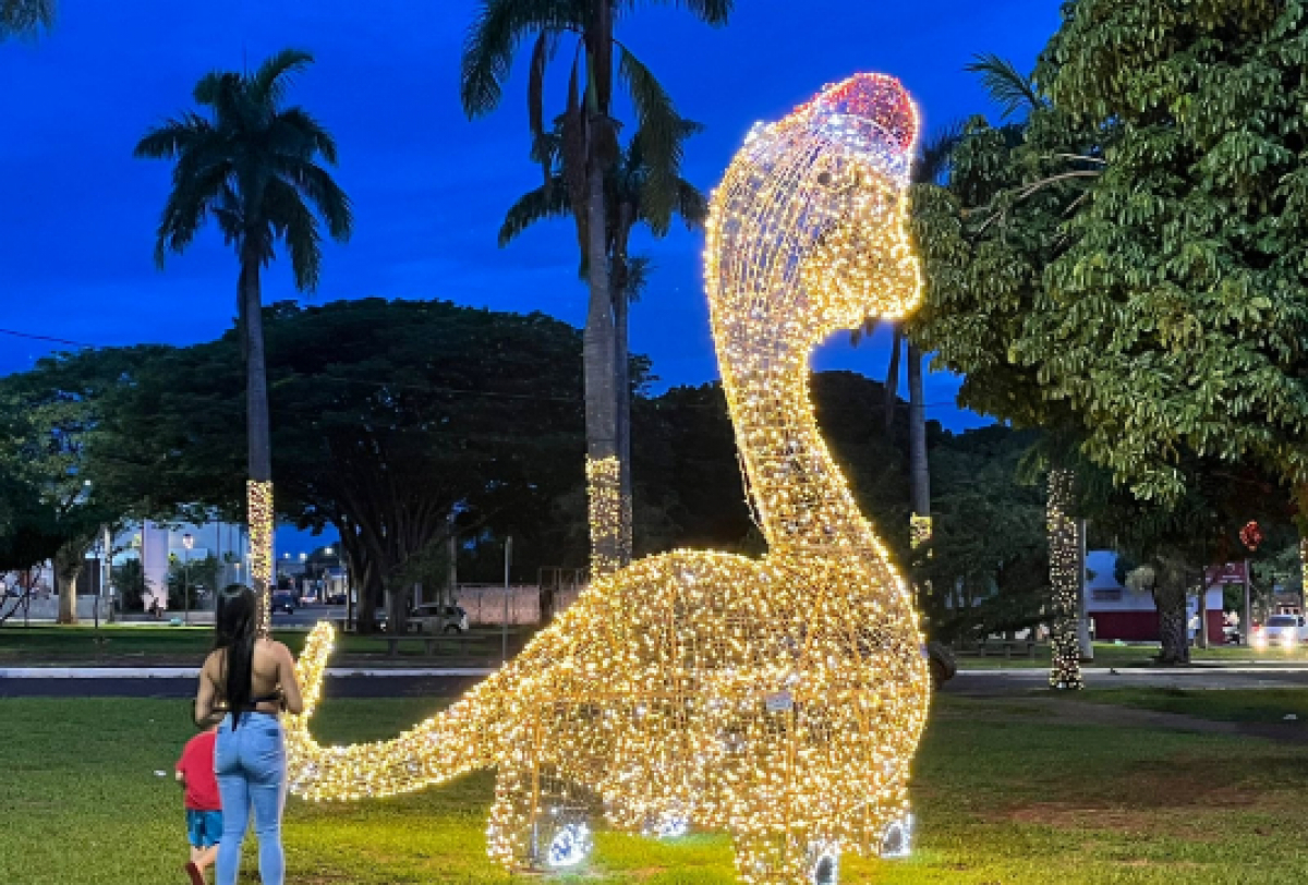 Instalação luminosa em formato de dinossauro embeleza praça ao anoitecer, criando um ambiente festivo.