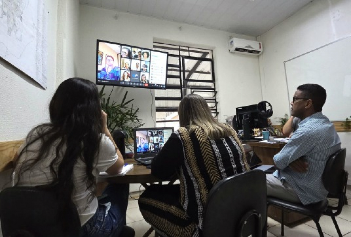 A imagem mostra um grupo de quatro pessoas em um pequeno escritório ou sala de reunião, participando de uma videoconferência.