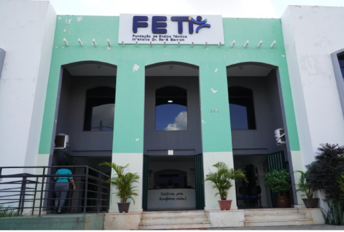 Fachada da FETI exibe entrada principal com plantas, visitantes e letreiro institucional.