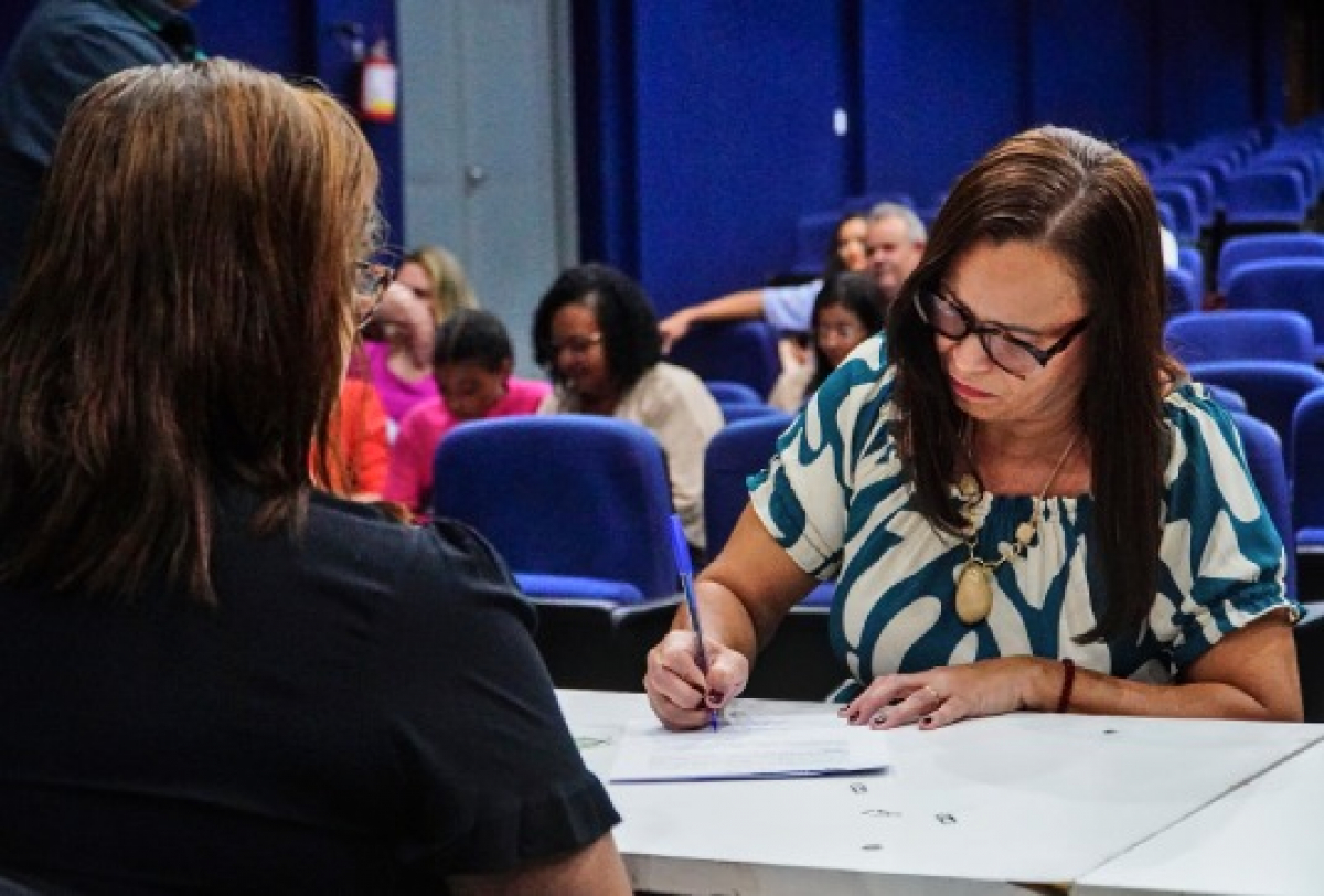Mulher preenche formulário durante atendimento em evento com várias pessoas em auditório.
