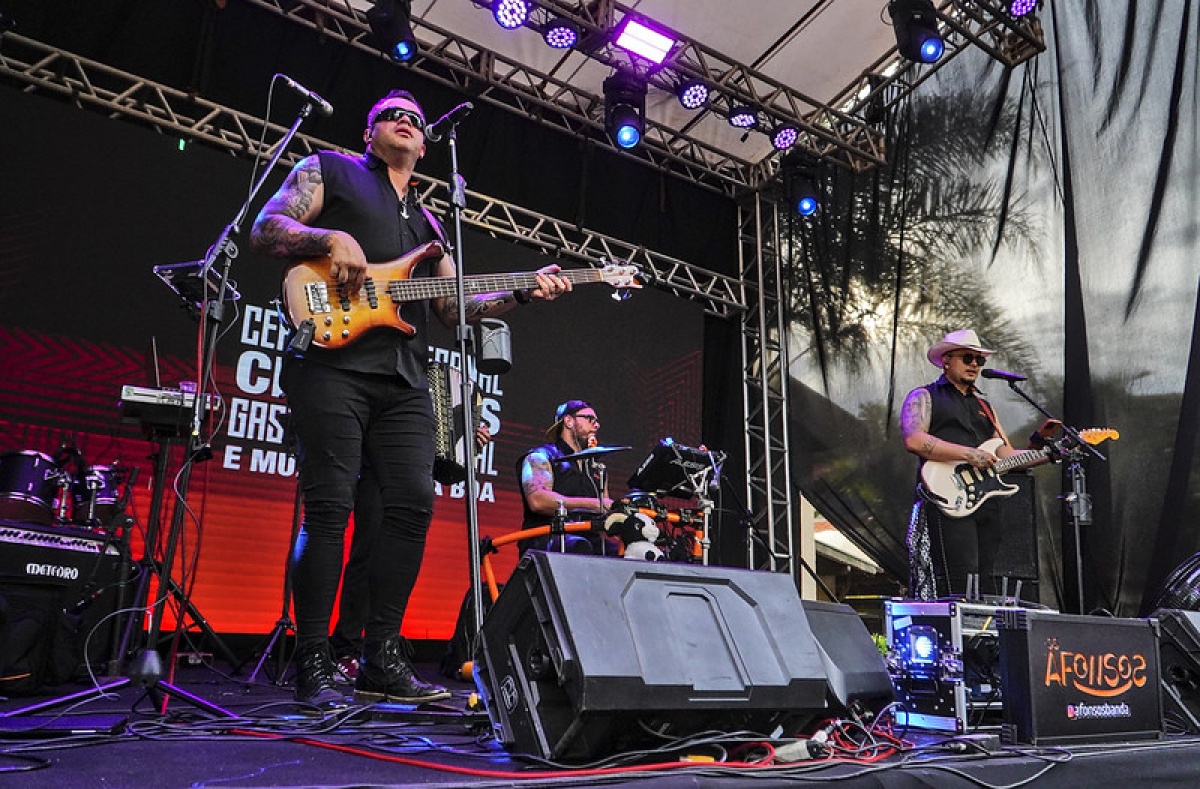 Banda se apresenta em palco ao ar livre durante evento musical, com guitarristas e baterista animando o público.